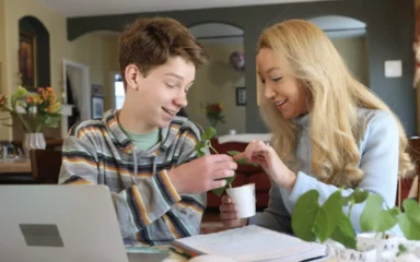 A young boy and a woman joyfully examine a small plant together at a table. The cozy room has warm lighting, flowers in the background, and an open laptop.