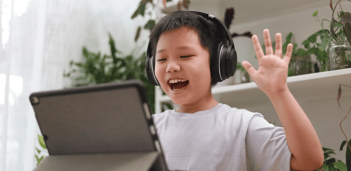 A cheerful child wearing headphones waves at a tablet screen during a video call at home, surrounded by green plants and soft natural light.