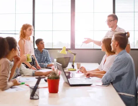 Group of six people in a bright office with large windows, engaged in discussion around a table with laptops and papers. A person stands, speaking animatedly.