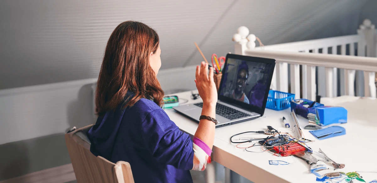 A woman sits at a desk waving at a laptop during a video call. The desk is cluttered with electronic components, suggesting a tech project setting.