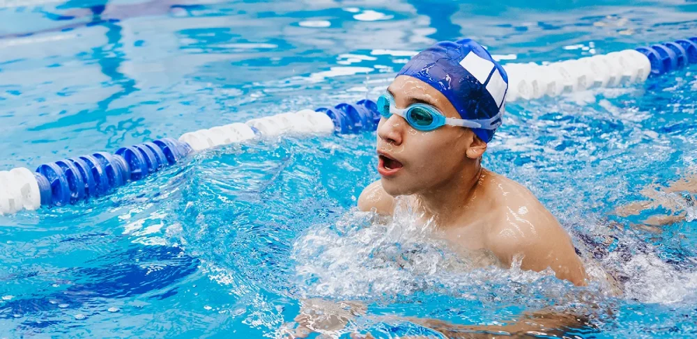 Young swimmer in blue cap and goggles doing breaststroke in pool, water splashing around him. Lane dividers visible, conveying focus and energy.
